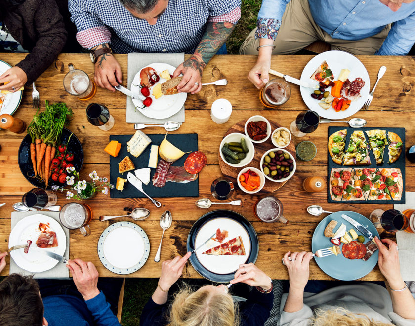 An overhead shot of people sitting around a farmhouse-style dinner table laid out with a bounty of food and drink